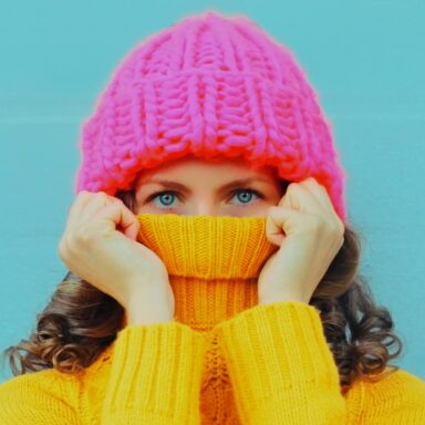 Woman in pink hat hiding the bottom of her face in her yellow jumper